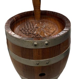 May include: A wooden barrel-shaped butter churn with metal bands. The top features a carved grape design and a wooden handle. The churn is made of dark brown wood and has a small hole near the bottom.