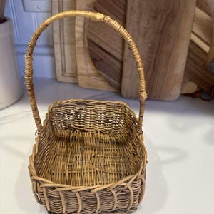 May include: A small, rectangular woven basket made of light brown natural reeds. The basket features a single, tall, arched handle attached to the sides, with a woven base and vertical reed walls that create a simple, open-top storage container.