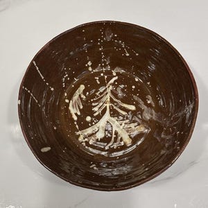 May include: A round, dark brown ceramic bowl with a glossy finish. The interior features a hand-painted, off-white abstract design resembling a stylized evergreen tree, with splattered paint accents extending toward the rim of the bowl.