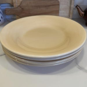 Pode incluir: Uma pilha de três pratos redondos de cerâmica em amarelo pálido com um acabamento liso e brilhante e uma borda ligeiramente elevada. Os pratos são uniformes em cor e forma, apresentando um design simples e limpo com um centro raso.