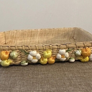 May include: Rectangular woven basket with a natural tan color. The basket is decorated with white, yellow, and orange floral embellishments along the bottom edge. The basket is likely used for storage or display.