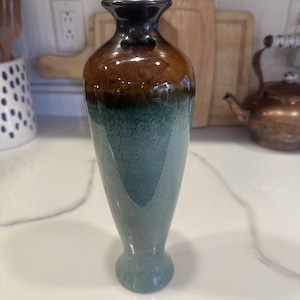 May include: A ceramic vase with a dark brown top, transitioning to a lighter brown, then teal, and ending in a teal base. The vase has a slender, elongated shape, suitable for displaying flowers or as a decorative piece. The vase is approximately 10 inches tall.