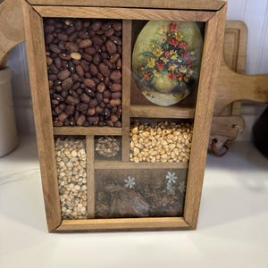 May include: A wooden wall hanging with a brass handle, featuring multiple compartments. The compartments contain beans, seeds, and a floral painting. The frame is a warm brown color, and the overall design is rustic and decorative.