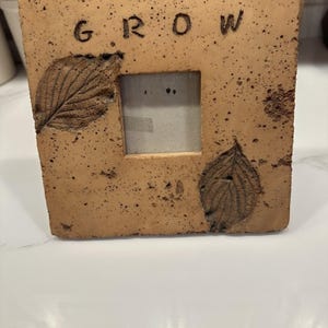Puede incluir: Un marco de fotos cuadrado de cerámica de color tostado con una textura moteada. La palabra GROW está estampada en letras negras en la parte superior. Dos impresiones de hojas oscuras en relieve son visibles a los lados de la abertura.