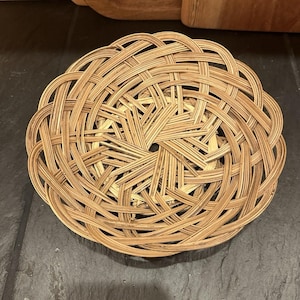 May include: A round, woven basket with an intricate, open weave design. The basket is crafted from light brown, natural-toned reeds, and features a circular form with a scalloped border. The basket rests on a dark grey surface.
