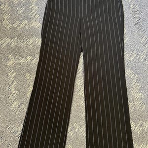 May include: Black wide-leg pants with vertical white pinstripes. The pants have a high waist and a wide leg opening. The fabric appears to be a medium-weight material, suitable for various occasions. The pants are laid flat on a patterned surface.