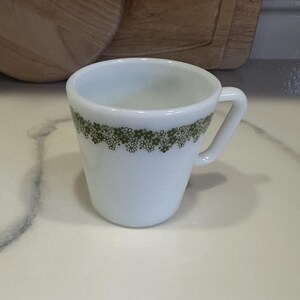 Puede incluir: Taza de café de cerámica blanca con un patrón floral verde alrededor del borde. La taza tiene un asa y una forma ligeramente acampanada. La taza está sobre una superficie blanca.