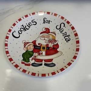 May include: A round white ceramic plate with a red and white striped rim. The center features a colorful illustration of Santa Claus holding two children, with the black text Cookies for Santa printed above. Small colorful confetti dots surround them.