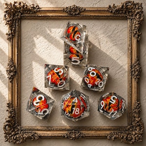 Könnte beinhalten: Set aus sieben Würfeln aus klarem Harz mit feurigen orangefarbenen und schwarzen Designs und weißen Zahlen. Die Würfel haben verschiedene geometrische Formen, darunter ein Würfel, ein d20 und ein d4, präsentiert in einem dekorativen goldenen Rahmen.