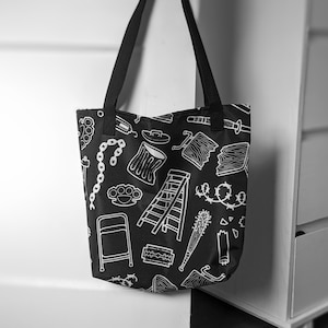 May include: Black tote bag with white line art illustrations of various objects, including a chain, brass knuckles, a ladder, and a barbed wire design. The bag has black handles and is hanging from a wooden knob.