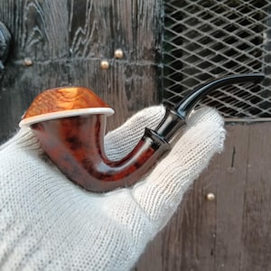Puede incluir: Una pipa de tabaco con un hornillo de madera pulida en color marrón oscuro con un patrón de veta distintivo, rematado con una tapa de madera de color marrón dorado más claro separada por un fino anillo blanco. Boquilla negra curva.