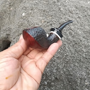 Può includere: Una pipa da tabacco marrone scuro con una testa arrotondata e un bocchino nero. La pipa ha una superficie strutturata sulla testa e una finitura liscia e lucida all'interno. Un anello bianco accentua la base del bocchino. La pipa è tenuta in mano su uno sfondo grigio.
