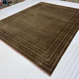 May include: A large rectangular area rug in shades of brown and tan, featuring a dense, repeating geometric pattern across the central field and a multi-layered border with intricate linear and wave-like motifs, finished with short fringe edges.