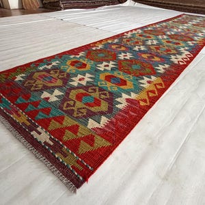 May include: A long, rectangular runner rug featuring a vibrant geometric pattern in shades of red, teal, yellow, and purple. The design consists of repeating diamond and triangular motifs with a fringed edge at the end of the woven textile.