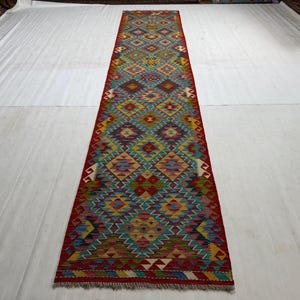 May include: A long, narrow rectangular rug featuring a vibrant, repeating geometric diamond pattern in shades of red, yellow, blue, green, and purple. The rug has a prominent red border and short, light-colored fringe at both ends of the piece.