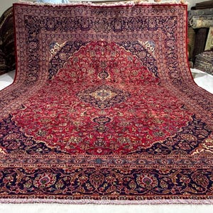 May include: A large rectangular area rug featuring a deep red central field with an intricate floral medallion and scrolling vine patterns in navy blue, cream, and gold. The wide border displays a complex navy blue floral motif with fringe.