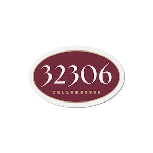 Puede incluir: Placa de dirección ovalada con borde blanco. El centro es burdeos con el número "32306" en escritura blanca. Debajo del número, la palabra "TALLAHASSEE" está impresa en una fuente más pequeña, sans-serif.