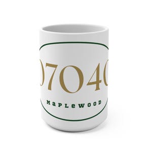 May include: White ceramic mug with the number "0704" in gold lettering, encircled by a green oval. The word "Maplewood" is written in green below the numbers. A simple, elegant design for a coffee or tea mug.