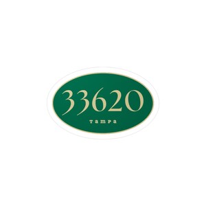 Puede incluir: Una pegatina ovalada con fondo verde oscuro y borde color crema. La pegatina presenta el número "33620" en una fuente estilizada, con la palabra "tampa" debajo en una fuente más pequeña.