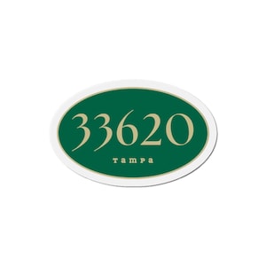 Puede incluir: Una pegatina ovalada con borde blanco, borde interior dorado y centro verde oscuro. El número "33620" es dorado, con "tampa" en dorado debajo. La pegatina es probablemente un identificador de código postal.