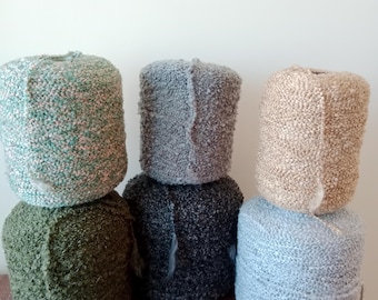 Puede incluir: Seis madejas de hilo en varios colores, incluyendo verde, gris y beige. El hilo tiene una apariencia texturizada y en bucle, lo que sugiere una textura suave y acogedora. Las madejas están apiladas en pares, mostrando las diferentes opciones de color.