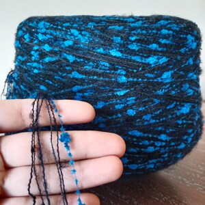 Könnte beinhalten: Nahaufnahme eines Wollknäuels, hauptsächlich schwarz mit leuchtend blauen Flecken. Fäden der Wolle werden in einer Hand gehalten, wodurch die Textur und die Farbvariationen hervorgehoben werden. Ideal für Strick- oder Häkelprojekte.