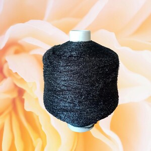 May include: A black yarn spool with a white base and top. The yarn has a fuzzy texture and is cylindrical in shape. The background is a soft, blurred peach color.