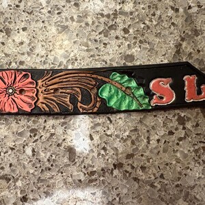 Op de afbeelding: Zwart leren riemgedeelte met bloemen- en bladeren tooling. Het ontwerp omvat een grote roze bloem, bruine bladeren en groene bladeren. De letters "SL" zijn rood. De riem is waarschijnlijk voor Western wear.