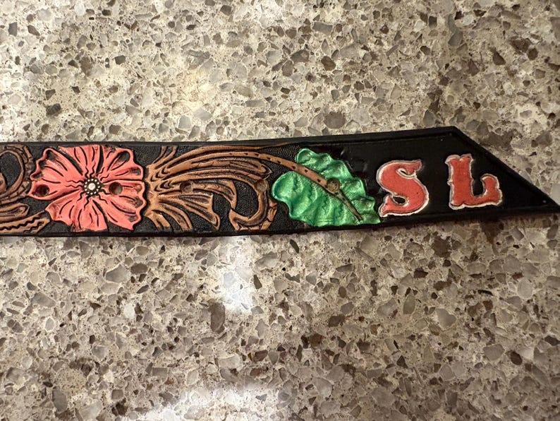 Op de afbeelding: Zwart leren riemgedeelte met bloemen- en bladeren tooling. Het ontwerp omvat een grote roze bloem, bruine bladeren en groene bladeren. De letters "SL" zijn rood. De riem is waarschijnlijk voor Western wear.