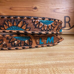 Op de afbeelding: Close-up van twee bruine leren riemen met zonnebloemontwerpen en turquoise accenten. De riemen hebben zwarte stiksels en een zilveren gesp. E&eacute;n riem heeft de initialen "LM" in turquoise.