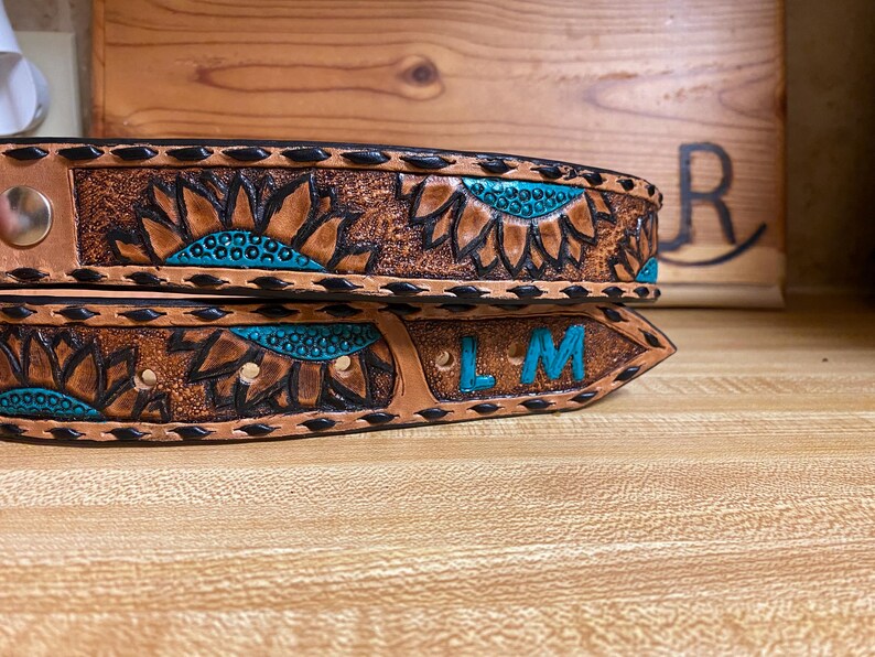 Op de afbeelding: Close-up van twee bruine leren riemen met zonnebloemontwerpen en turquoise accenten. De riemen hebben zwarte stiksels en een zilveren gesp. E&eacute;n riem heeft de initialen "LM" in turquoise.