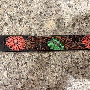 Op de afbeelding: Zwarte leren riem met reli&euml;f bloemmotief. De riem heeft drie koraalkleurige bloemen met witte centra, groene bladeren en bruine krullen. De riem heeft twee gaten en een gesp.