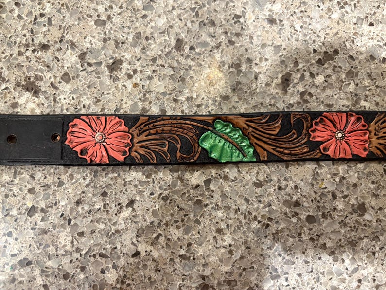 Op de afbeelding: Zwarte leren riem met reli&euml;f bloemmotief. De riem heeft drie koraalkleurige bloemen met witte centra, groene bladeren en bruine krullen. De riem heeft twee gaten en een gesp.