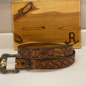 Op de afbeelding: Een bruine leren riem met bloemen en geometrische patronen. De gesp is zilverkleurig en draagt de letter "R". Op de achtergrond staat een houten bord met de letter "R".