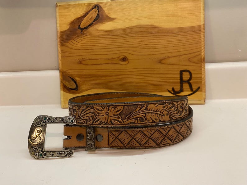 Op de afbeelding: Een bruine leren riem met bloemen en geometrische patronen. De gesp is zilverkleurig en draagt de letter "R". Op de achtergrond staat een houten bord met de letter "R".