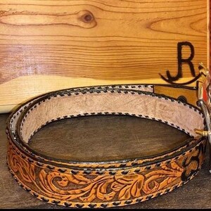 Op de afbeelding: Een bruine leren riem met ingewikkelde bloemenbewerking en een decoratieve zilveren gesp. De riem heeft een lichtbruin interieur en een donkerbruine rand met zwarte stiksels. De gesp heeft rode en oranje accenten.