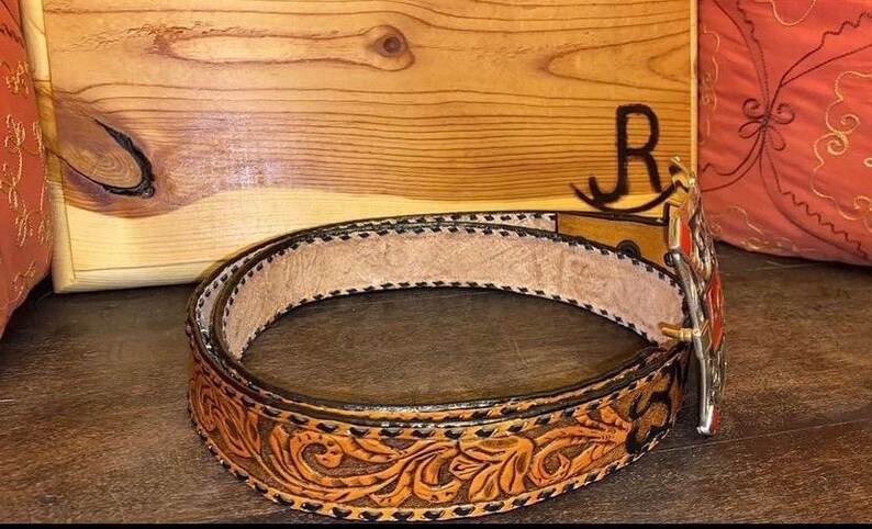Op de afbeelding: Een bruine leren riem met ingewikkelde bloemenbewerking en een decoratieve zilveren gesp. De riem heeft een lichtbruin interieur en een donkerbruine rand met zwarte stiksels. De gesp heeft rode en oranje accenten.