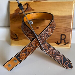 Op de afbeelding: Een bruine leren gitaarband met bloemmotieven en zwarte randen, gekruist over een houten achtergrond. Een metalen plectrumhouder is door de band gestoken. De initialen "CL" zijn zichtbaar op de band.
