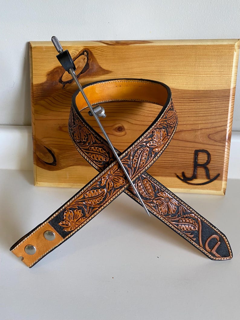 Op de afbeelding: Een bruine leren gitaarband met bloemmotieven en zwarte randen, gekruist over een houten achtergrond. Een metalen plectrumhouder is door de band gestoken. De initialen "CL" zijn zichtbaar op de band.