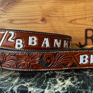 Op de afbeelding: Een bruine leren riem met witte letters met de tekst "1/2 B BANKS" en "BB". De riem heeft bloemen- en krulontwerpen. De riem ligt op een donkere ondergrond.