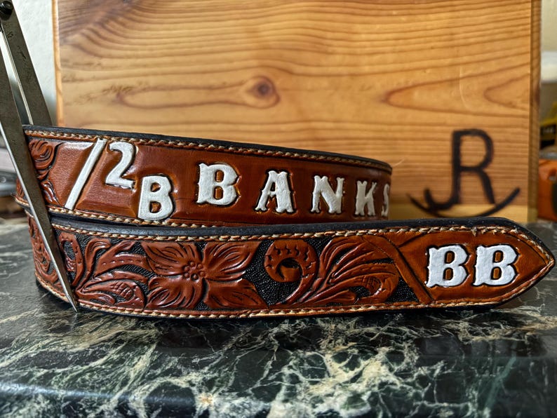 Op de afbeelding: Een bruine leren riem met witte letters met de tekst "1/2 B BANKS" en "BB". De riem heeft bloemen- en krulontwerpen. De riem ligt op een donkere ondergrond.