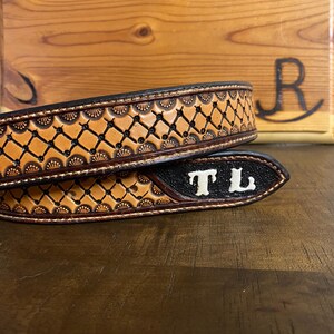 Op de afbeelding: Een close-up van een bruine leren riem met ingewikkelde bewerkingen. De riem heeft een herhalend geometrisch patroon en is gepersonaliseerd met de initialen "TL" in wit. De achtergrond omvat een houten oppervlak en een houten bord.