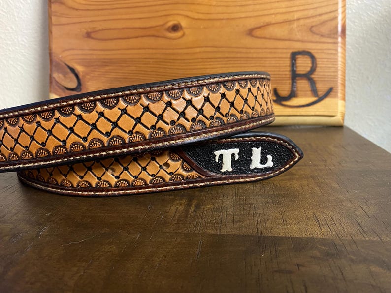 Op de afbeelding: Een close-up van een bruine leren riem met ingewikkelde bewerkingen. De riem heeft een herhalend geometrisch patroon en is gepersonaliseerd met de initialen "TL" in wit. De achtergrond omvat een houten oppervlak en een houten bord.