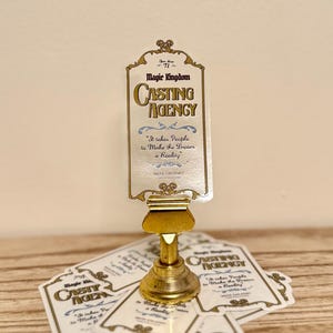 May include: A gold-colored Magic Kingdom Casting Agency sign with the text "It takes People to Make the Dream a Reality." The sign is on a gold stand, with additional signs scattered below.