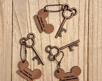 Könnte beinhalten: Drei schlüsselförmige Flaschenöffner in Bronze mit einem Mickey Mouse-Kopf-Design. Jeder Öffner ist mit einem Holzanhänger mit der Aufschrift "Thank You For Making Magic" versehen. Sie liegen auf einer Holzoberfläche.