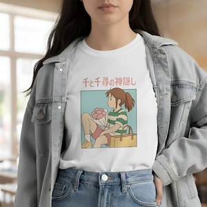 Puede incluir: Camiseta blanca con una ilustraci&oacute;n de estilo anime de una ni&ntilde;a sosteniendo flores y una bolsa. La ni&ntilde;a lleva una camiseta a rayas verdes y blancas y pantalones cortos rojos. Texto japon&eacute;s encima de la ilustraci&oacute;n.