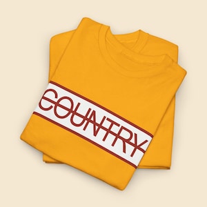May include: Two mustard yellow t-shirts are stacked. The top shirt features the word "COUNTRY" in white letters on a diagonal red-bordered white stripe. The shirts are neatly folded, highlighting the design.