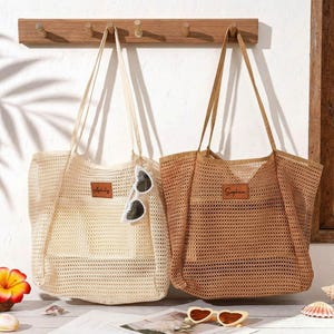 May include: Two mesh tote bags, one cream and one tan, hang from a wooden rack. Each bag has a rectangular leather patch with a name on it. Sunglasses and beach-themed decor are in the foreground, with a beach scene visible in the background.