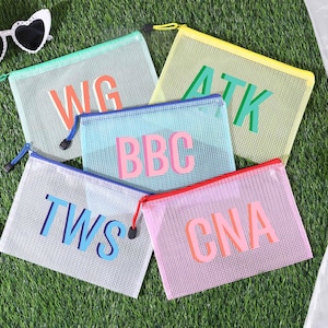 May include: Five mesh zipper pouches in various colors, each with a different set of three capital letters. The pouches are on a green surface, with sunglasses and a hat in the background. The pouches have colored zippers.