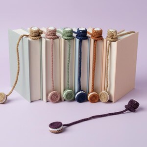 May include: A collection of handmade crochet bookmarks in various colors, including brown, pink, green, blue, and tan. Each bookmark features a decorative top and a matching circular bottom, resting on a stack of books. One bookmark lies separately.